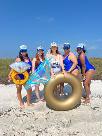 Five women in matching blue swimsuits and trucker hats posing on a sunny sandy beach for a bachelorette celebration, holding a gold inner tube and oversized diamond-ring inflatables.