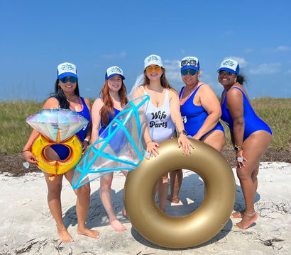 Five women in matching blue swimsuits and trucker hats posing on a sunny sandy beach for a bachelorette celebration, holding a gold inner tube and oversized diamond-ring inflatables.