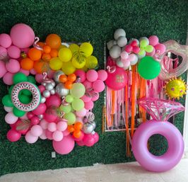 Bright tropical party backdrop on a faux green hedge wall with a multicolor balloon garland (pink, orange, yellow, green, silver), inflatable watermelon float, oversized pink diamond-ring float, smiling sun balloon, flamingo and pineapple accents, and pink fringe streamers.