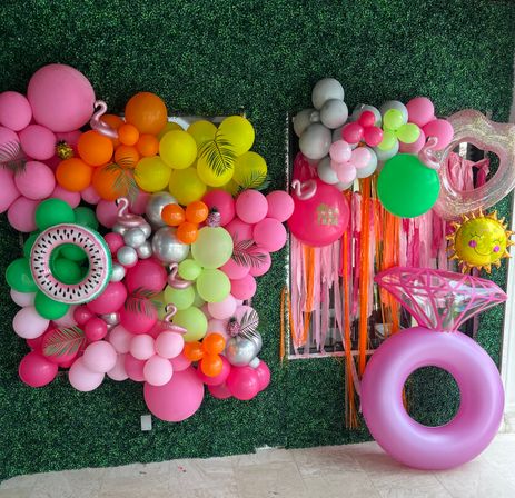 Bright tropical party backdrop on a faux green hedge wall with a multicolor balloon garland (pink, orange, yellow, green, silver), inflatable watermelon float, oversized pink diamond-ring float, smiling sun balloon, flamingo and pineapple accents, and pink fringe streamers.