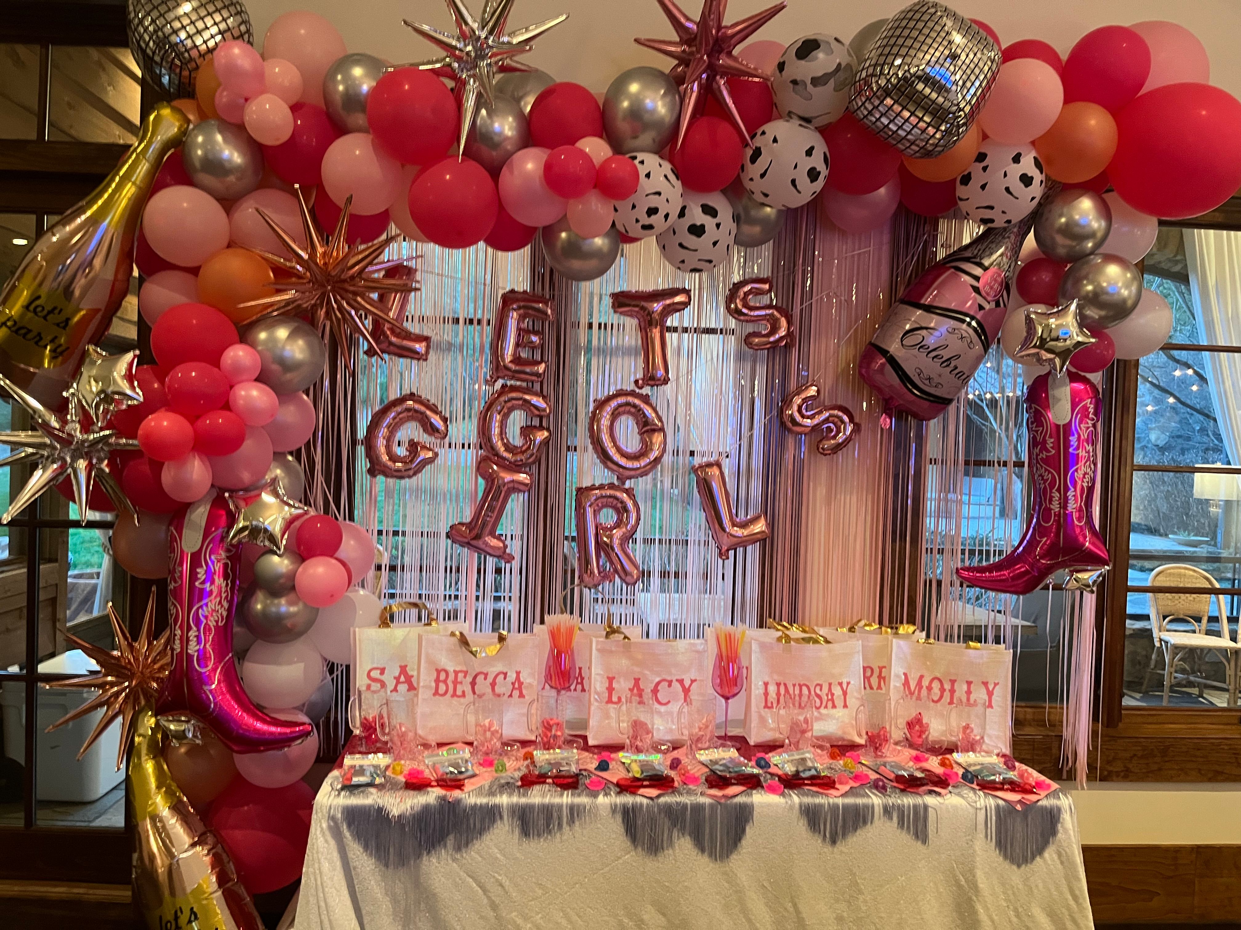 Indoor pink-and-metallic balloon arch and shimmering fringe backdrop with foil letter balloons spelling LET'S GO GIRLS, star and disco-ball accents, champagne and cowboy-boot foil balloons, and a table of white favor bags and pink party treats.