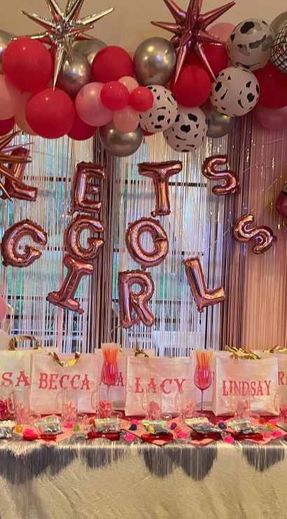 Indoor pink-and-metallic balloon arch and shimmering fringe backdrop with foil letter balloons spelling LET'S GO GIRLS, star and disco-ball accents, champagne and cowboy-boot foil balloons, and a table of white favor bags and pink party treats.