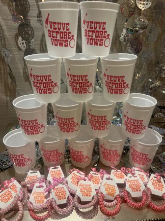 Pyramid of white plastic cups printed “Veuve Before Vows” in pink with ring and champagne icons, displayed above pink bachelorette wristband favors on a sparkly backdrop.