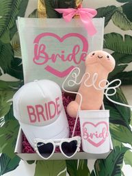 Pink-and-white bachelorette gift box for bride with 'BRIDE' trucker hat, heart sunglasses, plush novelty toy, curly-straw cup and matching tote bag