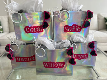 Five iridescent party-favor bags with white tissue paper and pink heart-shaped sunglasses clipped to each, pink name labels visible, arranged on a glass coffee table in a bright living room.