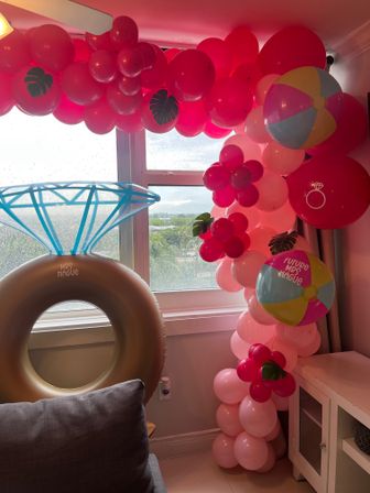 Playful pink balloon garland and flower clusters framing an ocean-view window, with a large inflatable diamond-ring float and colorful beach-ball balloons for a bridal shower celebration.