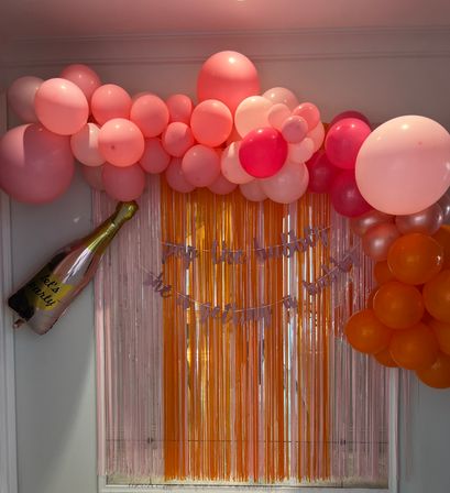 Cheerful pink and coral balloon garland arching over orange and pink metallic fringe photo backdrop with a foil champagne bottle balloon — indoor party decoration