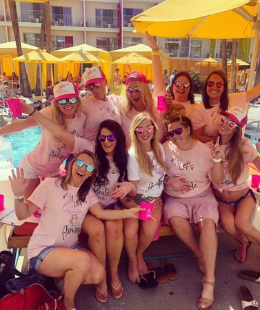 Poolside bachelorette crew in matching pink flamingo tees and mirrored sunglasses, cheering with pink cups under yellow umbrellas at a sunny hotel pool