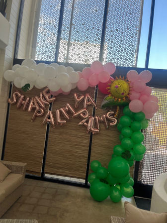 Indoor tropical-themed party backdrop with white and pink balloon clouds, a green balloon palm column and smiling sun foil balloon, rose-gold letter balloons on a wood-paneled wall beneath tall patterned windows, lounge seating visible.