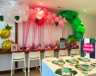 Indoor tropical-themed party setup with pink and green balloon garland and streamer backdrop, pineapple and sun foil balloons, white bar stools, decorated snack table with party favors and a 'Press for Champagne' sign.
