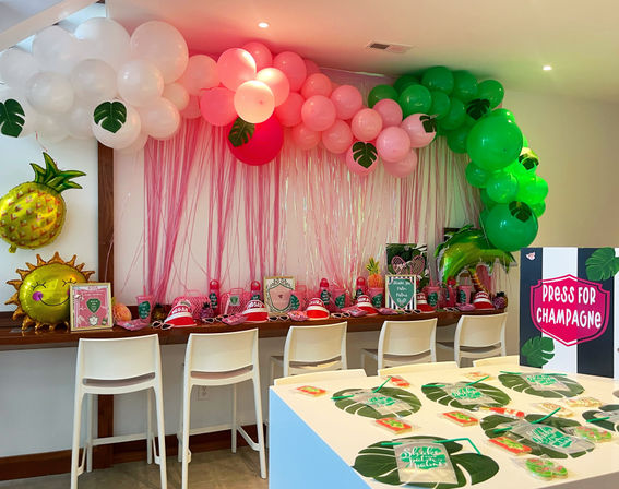 Indoor tropical-themed party setup with pink and green balloon garland and streamer backdrop, pineapple and sun foil balloons, white bar stools, decorated snack table with party favors and a 'Press for Champagne' sign.