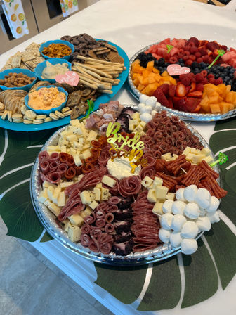 Colorful bridal-shower appetizer spread on a kitchen island: large charcuterie platter with rolled cured meats, cubes of cheese, mozzarella balls and a 'Bride to be' topper, nearby cracker and dip board, and a fresh fruit tray of strawberries, raspberries, blueberries and cantaloupe.