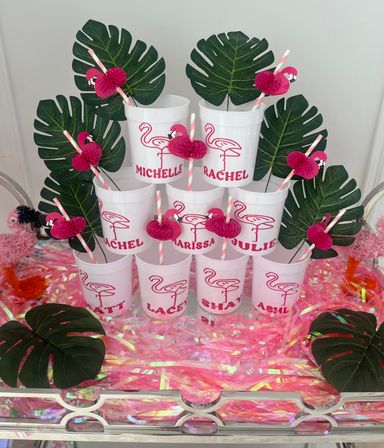 Tropical flamingo party display of stacked white cups with pink flamingo graphics and personalized names, striped straws topped with honeycomb pink flamingos, large faux monstera leaves and iridescent pink fringe.