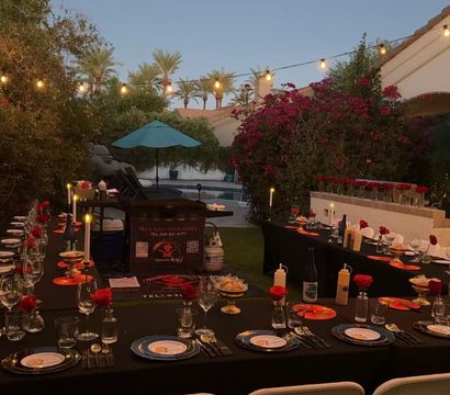 Twilight poolside backyard dinner with long black-tablecloth tables, formal place settings, red roses, candles and string lights under palm trees and bougainvillea