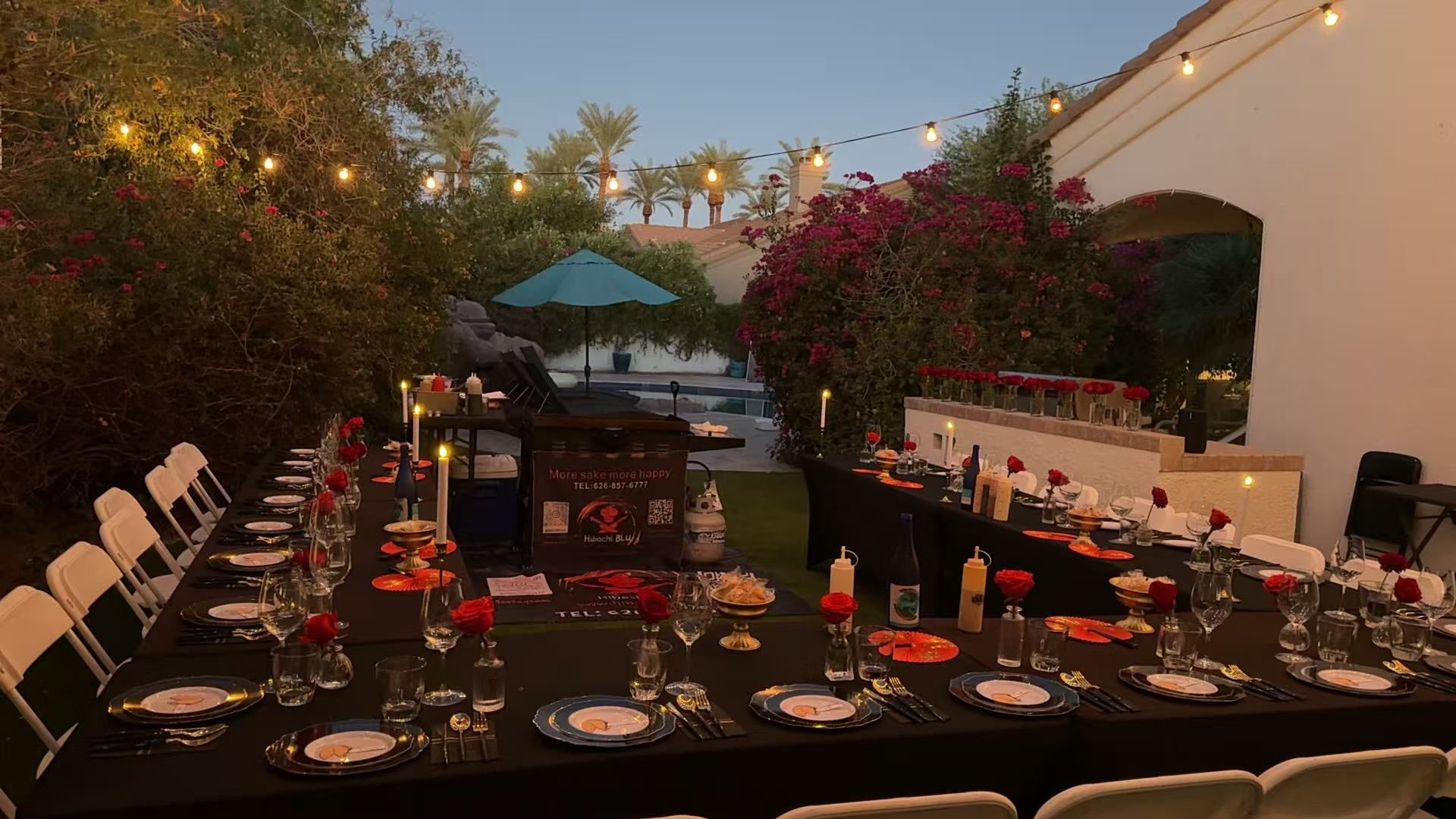 Twilight poolside backyard dinner with long black-tablecloth tables, formal place settings, red roses, candles and string lights under palm trees and bougainvillea