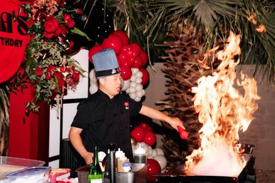 Chef in blue hat creating a dramatic flaming teppanyaki-style flambé at an outdoor red-and-white balloon party with roses and palm fronds