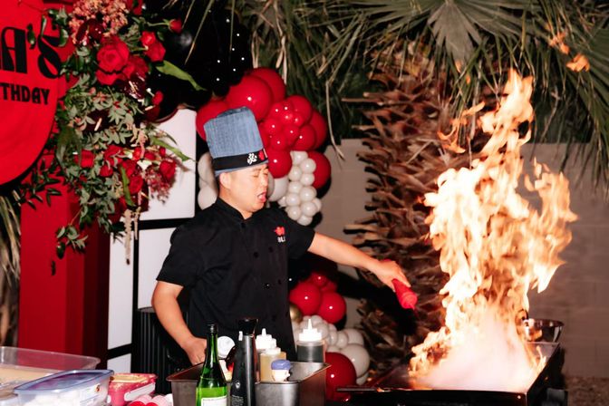 Chef in blue hat creating a dramatic flaming teppanyaki-style flambé at an outdoor red-and-white balloon party with roses and palm fronds