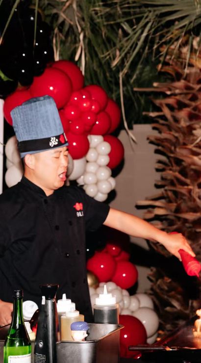 Chef in blue hat creating a dramatic flaming teppanyaki-style flambé at an outdoor red-and-white balloon party with roses and palm fronds