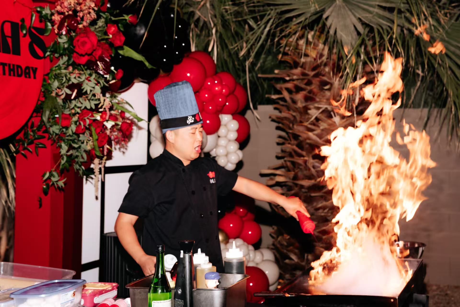Chef in blue hat creating a dramatic flaming teppanyaki-style flambé at an outdoor red-and-white balloon party with roses and palm fronds