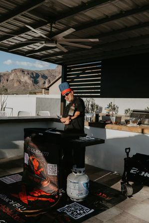 Outdoor patio kitchen with a cook in a blue chef hat flipping food on a flat-top griddle under a large ceiling fan, propane tank and prep counter visible, rugged desert mountains beyond.