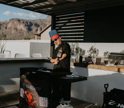 Outdoor patio kitchen with a cook in a blue chef hat flipping food on a flat-top griddle under a large ceiling fan, propane tank and prep counter visible, rugged desert mountains beyond.