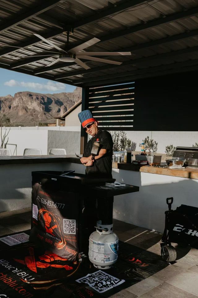 Outdoor patio kitchen with a cook in a blue chef hat flipping food on a flat-top griddle under a large ceiling fan, propane tank and prep counter visible, rugged desert mountains beyond.