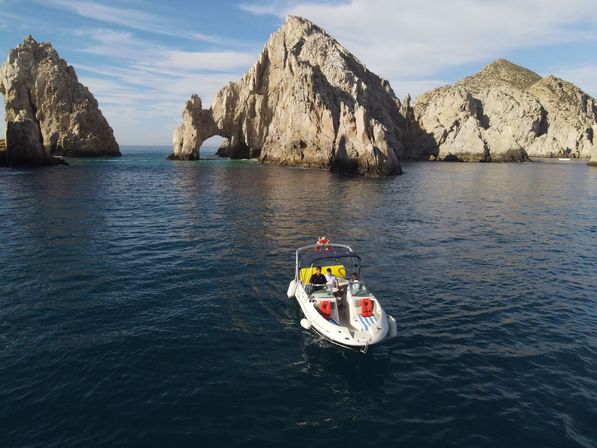 Small white motorboat with passengers on deep blue ocean in front of a dramatic coastal rock arch and rugged cliffs under a sunny sky