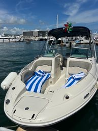 White motorboat with blue-and-white striped beach towels on the bow, black bimini top and a colorful pinwheel, docked at a sunny marina with yachts and waterfront buildings