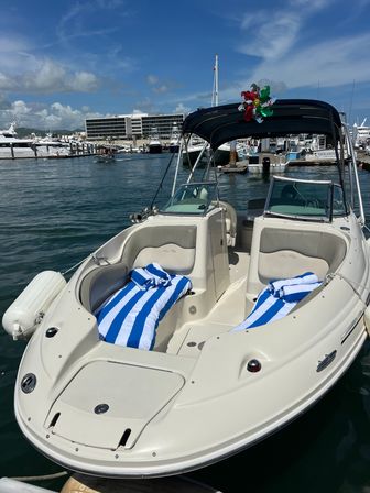 White motorboat with blue-and-white striped beach towels on the bow, black bimini top and a colorful pinwheel, docked at a sunny marina with yachts and waterfront buildings