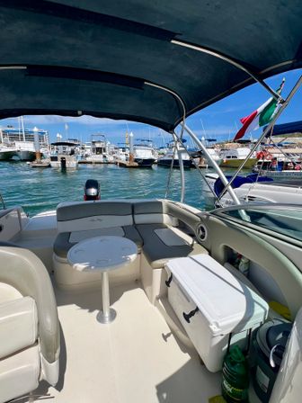 Sunlit motorboat cockpit with curved bench seating, small round table and cooler, overlooking a busy marina in Mexico with docked boats and a Mexican flag.