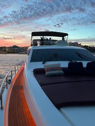 Sunset yacht bow with cushioned sunpad and patterned pillow, warm-lit teak side deck, silhouetted people on the flybridge and a pastel sky over a coastal harbor.