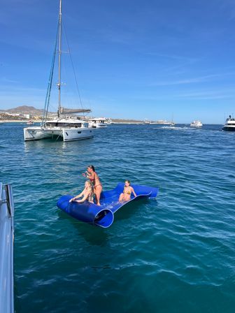 Three people in swimsuits on a bright blue inflatable floating mat near anchored catamarans and yachts in clear turquoise ocean under a sunny sky in a coastal bay