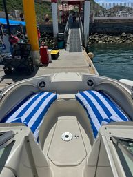 Bow view of a leisure motorboat with blue-and-white striped towels on cushioned seats, docked at a sunny marina ramp beside calm harbor waters and rocky shoreline.