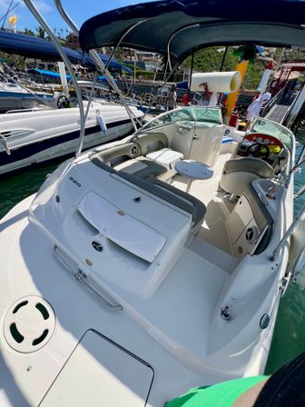 White recreational motorboat with cushioned seating, small round table and navy bimini top docked in a sunny coastal marina beside other boats over clear green water.