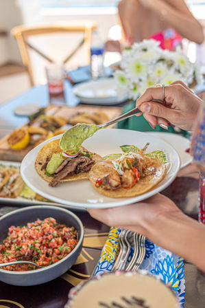 Hand spooning fresh guacamole onto two street-style tacos—steak and shrimp—on a white plate with pico de gallo, grilled lemons and a colorful table setting for a lively Mexican-style brunch.