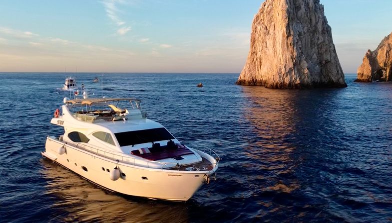 White luxury yacht gliding past a towering rocky sea stack at sunset, calm blue ocean reflecting warm golden light