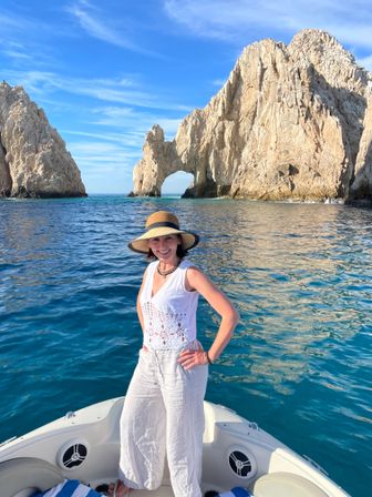 Woman in a straw hat on a boat tour in front of the El Arco rock arch at Cabo San Lucas, Mexico, with turquoise sea and clear blue sky.