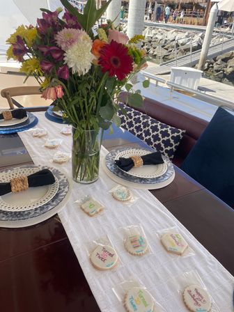 Yacht dining table at a marina with a tall vase of mixed bright flowers, blue-patterned plates and woven napkin rings, and individually wrapped birthday cookies on a white runner.