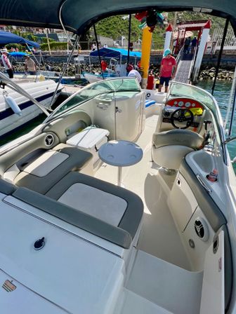 Sunny coastal motorboat cockpit with gray cushioned seating, small round table and helm, docked at a busy marina pier