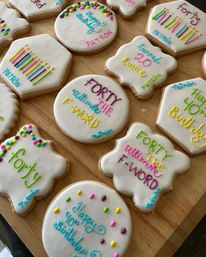 Assorted hand-decorated birthday sugar cookies with white royal icing and colorful piped lettering, dots and candle designs featuring 40th birthday messages, arranged on a wooden board.