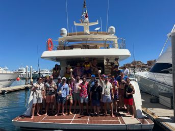 Group enjoying a yacht party on the stern of a luxury boat docked in a sunny marina, wearing summer outfits and leis, holding tropical drinks with clear blue sky and other yachts in the background