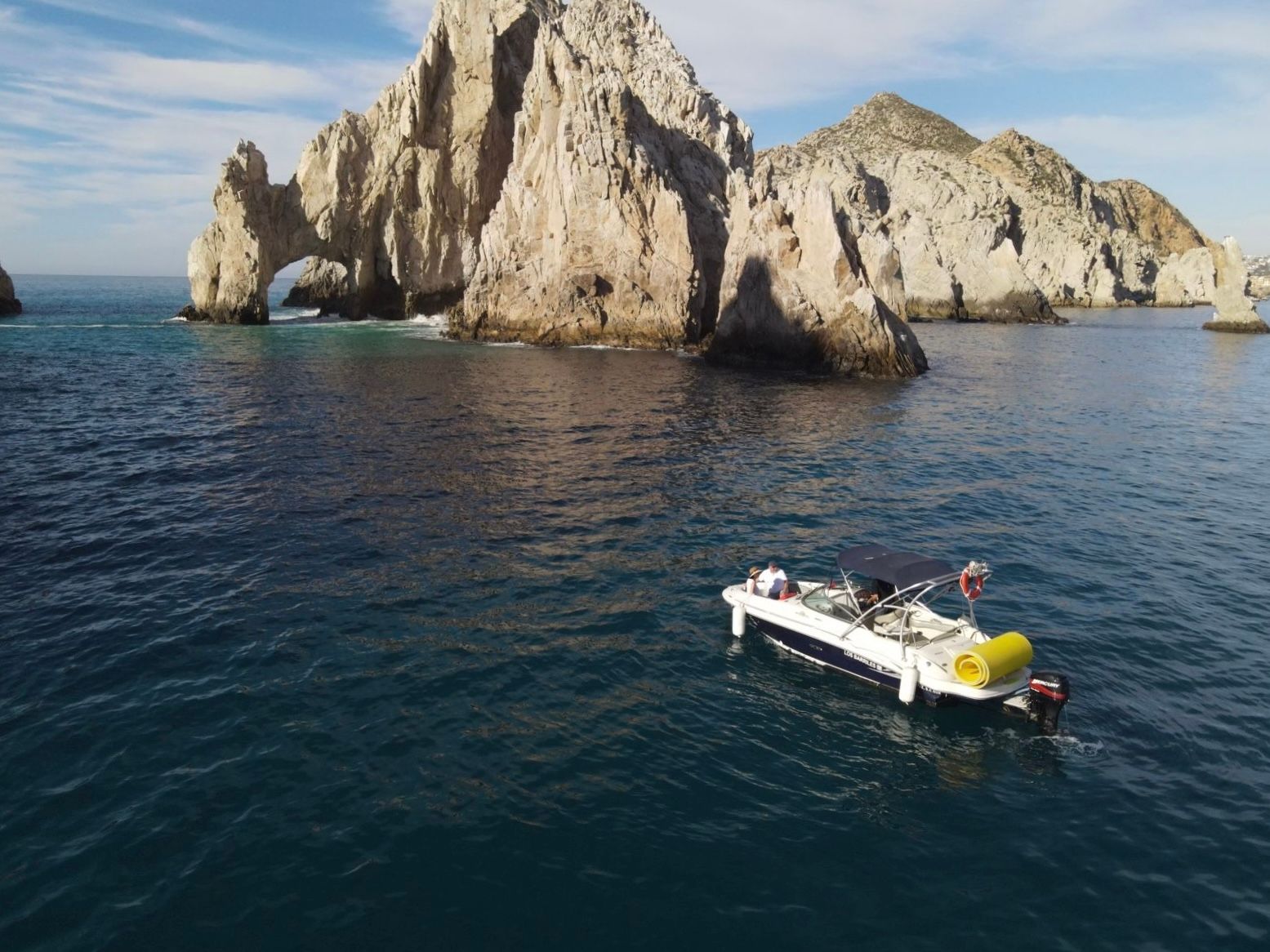 Small motorboat floating in deep blue ocean near a dramatic coastal rock arch and sunlit limestone cliffs under a clear sky.