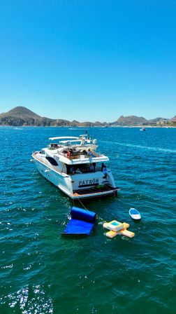 Sun-soaked white luxury yacht anchored near a rocky coastline under a clear blue sky, turquoise water with a blue floating sun mat, inflatable lounge and paddleboard.