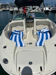 Bow of a white motorboat docked at a sunny marina, twin lounge seats with blue-and-white striped beach towels, windshield and bimini shade, other boats and harbor buildings in the background.
