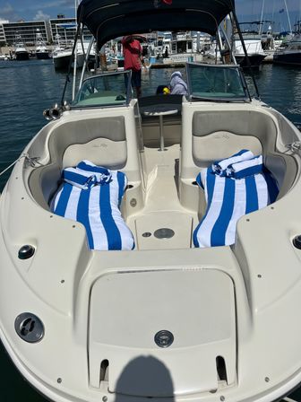 Bow of a white motorboat docked at a sunny marina, twin lounge seats with blue-and-white striped beach towels, windshield and bimini shade, other boats and harbor buildings in the background.