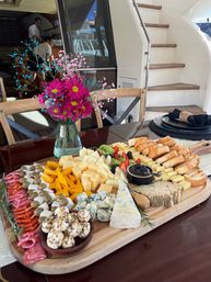 Vibrant charcuterie and cheese board with assorted cheeses, cured meats, marinated cheese balls, fresh berries and sliced baguette on a wood table beside bright pink flowers aboard a yacht at the marina.
