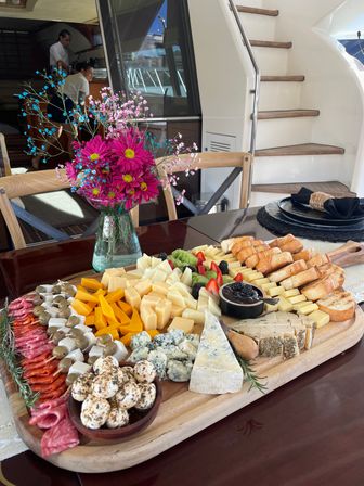 Vibrant charcuterie and cheese board with assorted cheeses, cured meats, marinated cheese balls, fresh berries and sliced baguette on a wood table beside bright pink flowers aboard a yacht at the marina.