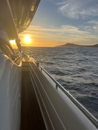 Golden-hour sunset view from a yacht side deck, stainless railings and teak walkway overlooking choppy ocean waters toward a distant coastal silhouette.