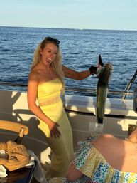 Sunlit woman in a yellow sundress on a boat holding up a freshly caught fish with a gloved hand, smiling against the open ocean horizon.