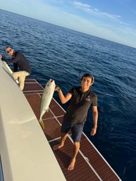 Two smiling men on a boat in the deep blue ocean during an offshore fishing trip — one holding a large catch over the teak swim platform under a clear sunny sky.
